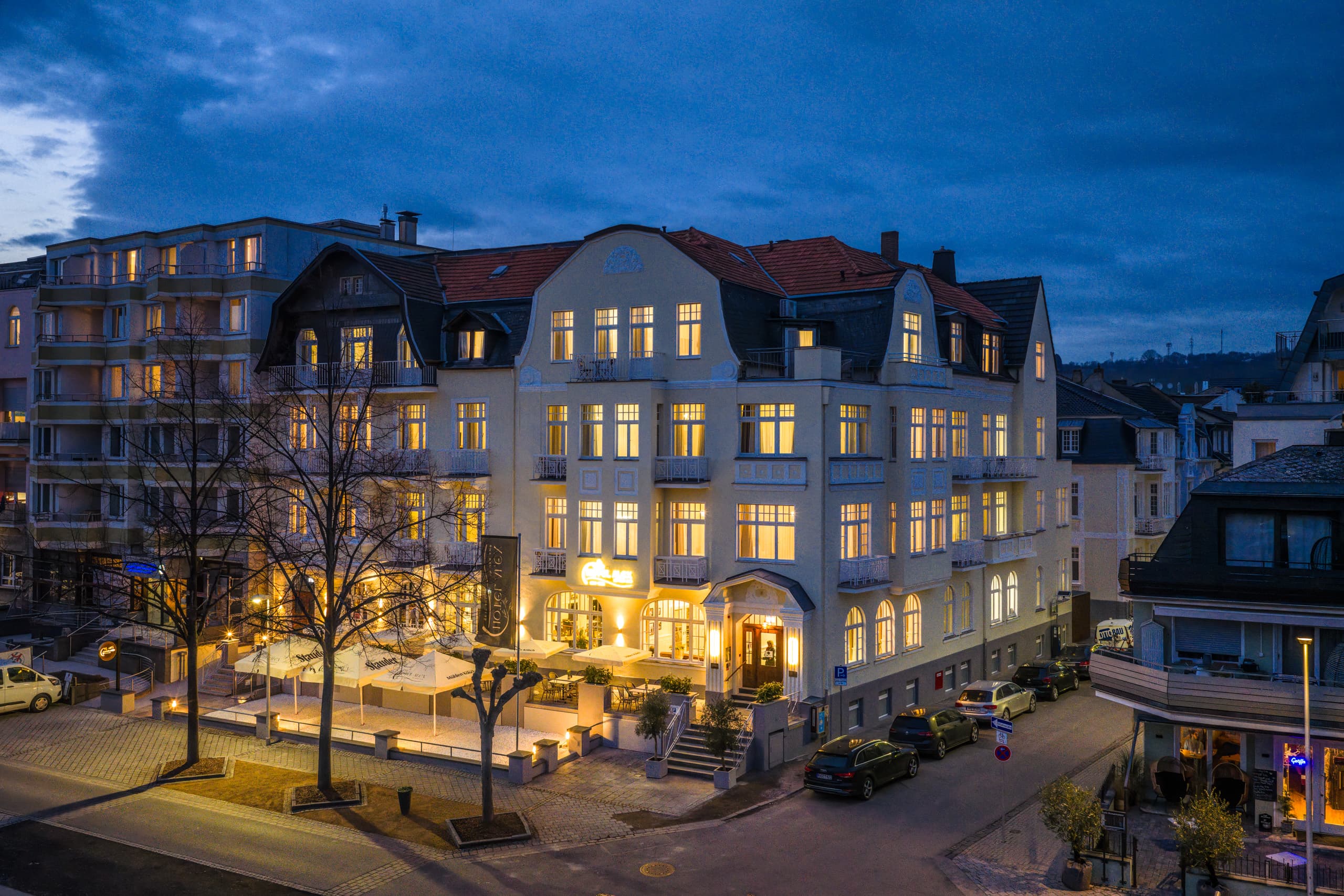 Die beleuchtete Jugendstilfassade des Hotel Alex mit Terrasse in der Abenddämmerung
