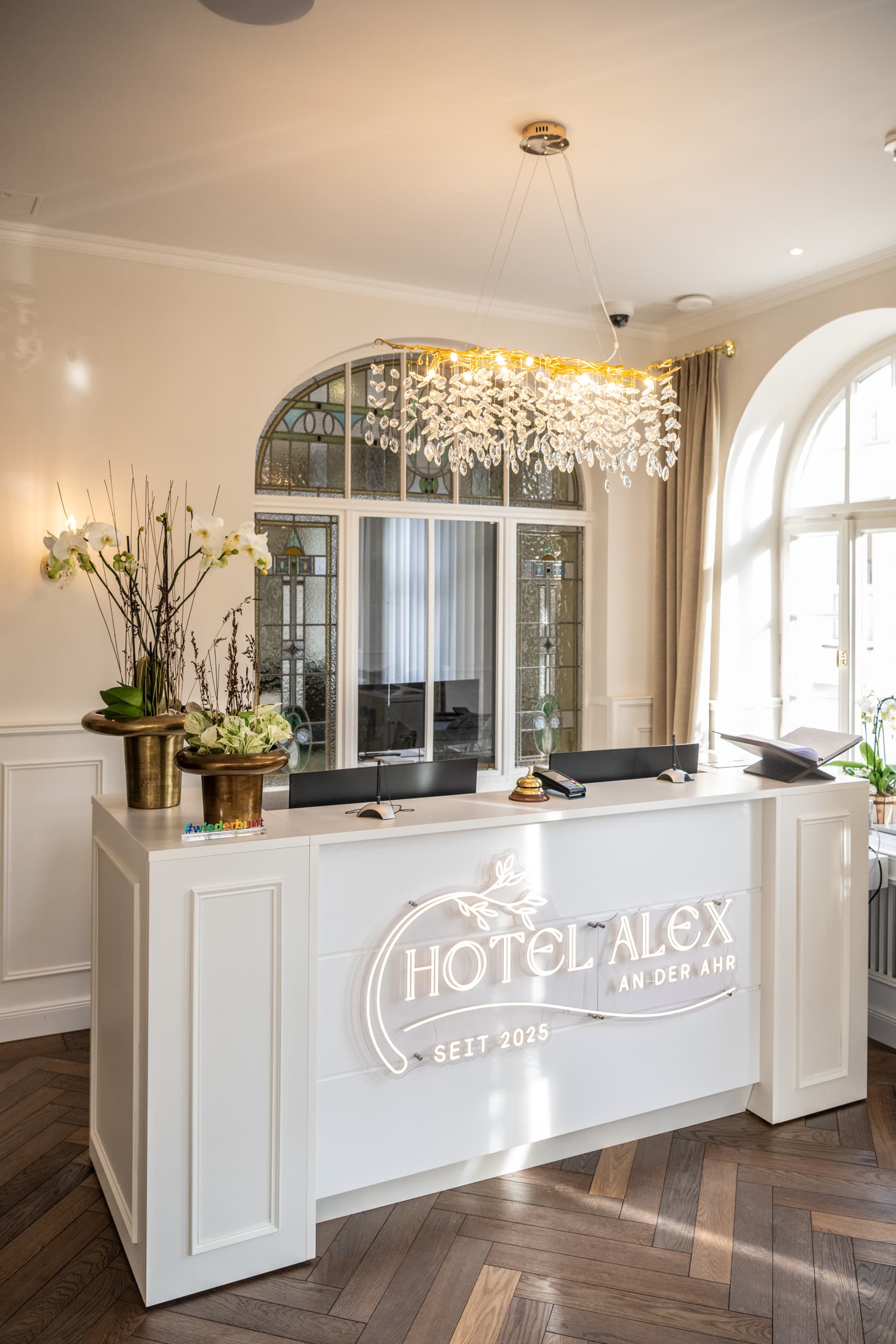 Weiße Rezeption mit Hotel-Alex-Schriftzug, Kronleuchter und Buntglasfenster