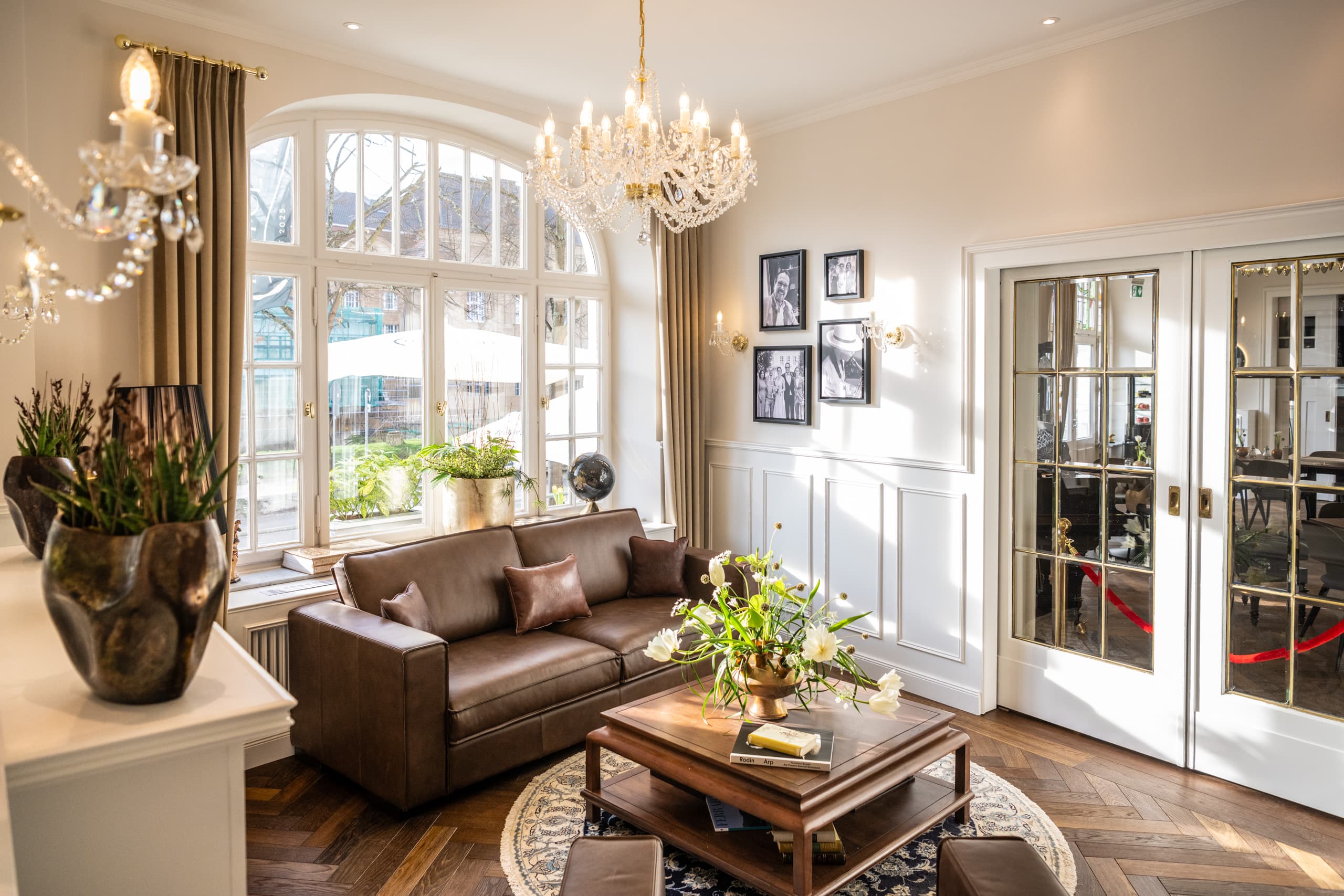 Foyer mit Ledersofas, Blumen und Kronleuchter vor großen Rundbogenfenstern