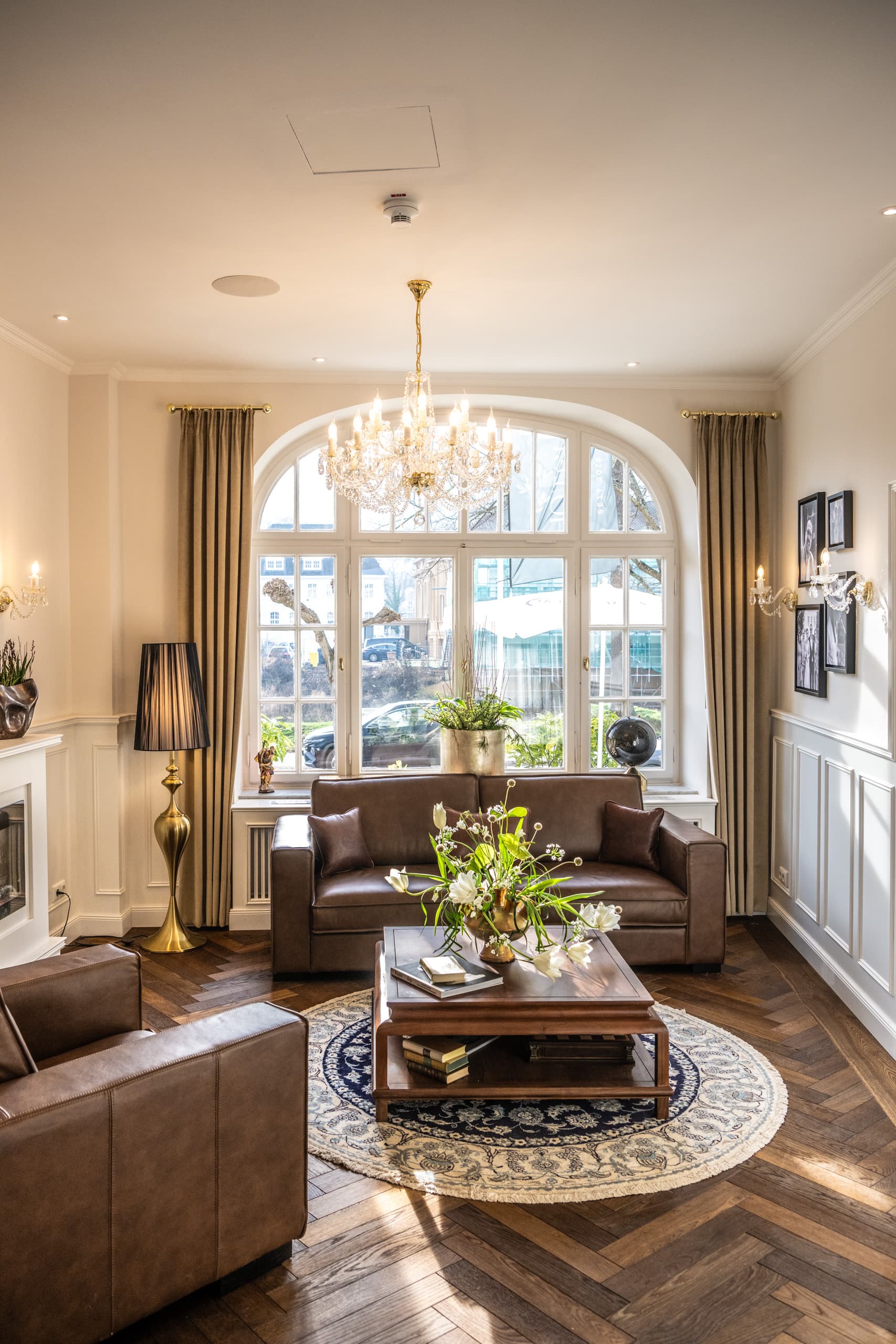 Foyer mit braunen Ledersesseln, Sofa und Kronleuchter vor dem Rundbogenfenster